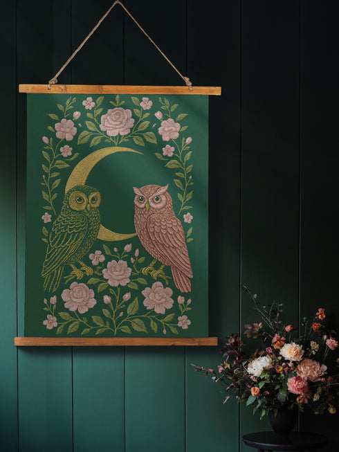 26x36 inch vertical wall tapestry printed on silky polyester, featuring a pair of embroidered-style owls perched on a golden crescent moon, surrounded by pink vintage-style florals and greenery on a forest green background. Includes stained wood slats and twine for easy hanging. A romantic, nature-inspired design for woodland and cottagecore decor lovers.